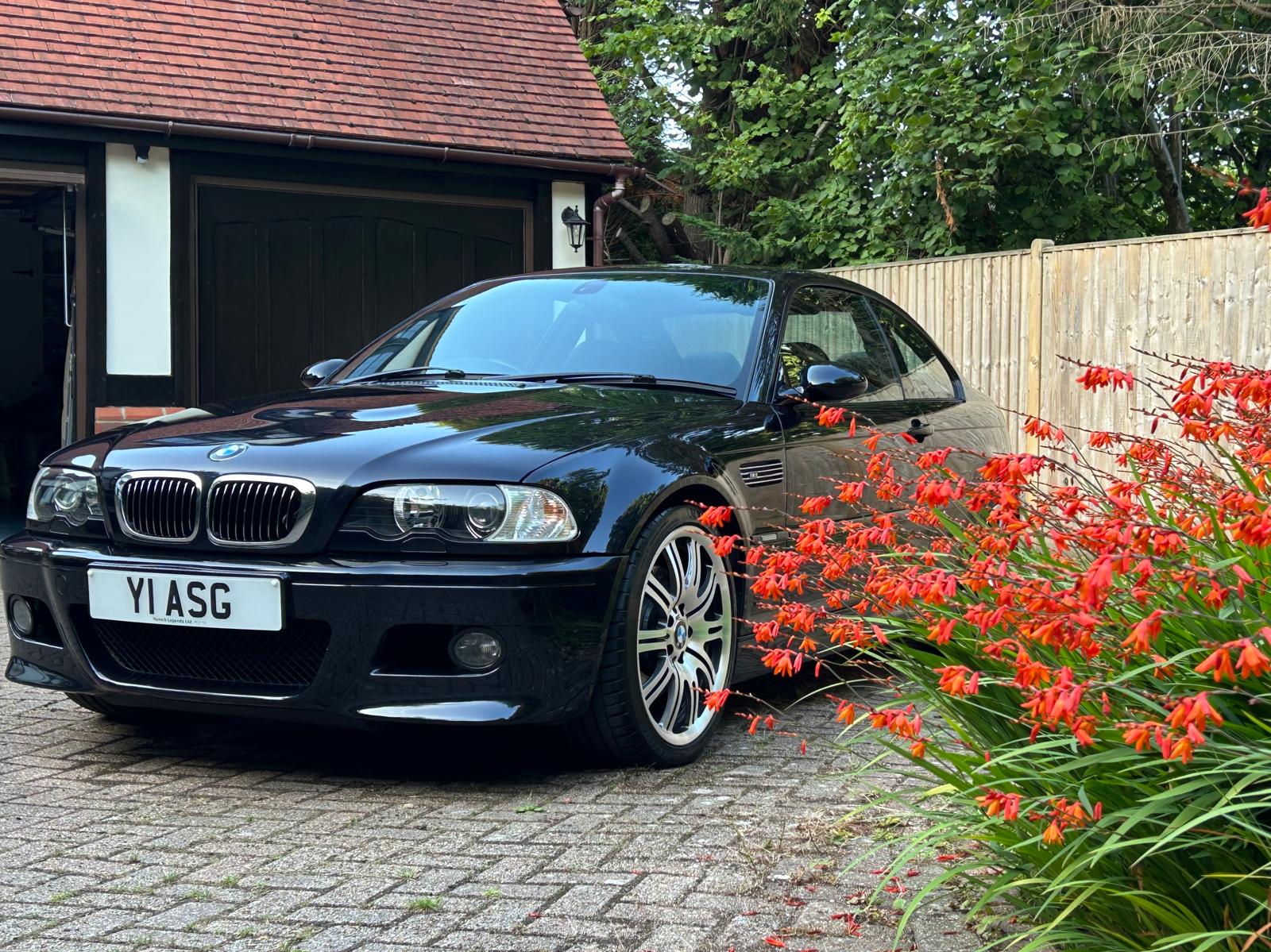 Black BMW E46 M3 coupe parked beside red flowers on a driveway